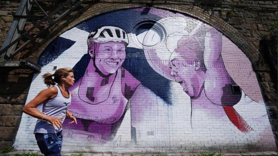 A runner runs past a white, purple and blue mural featuring Team Scotland stars.