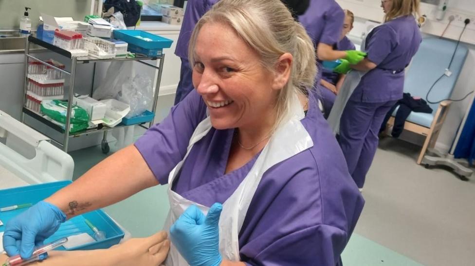 Rachel wearing purple hospital scrubs and blue surgical gloves. She smiles at the camera and holds a syringe in one hand. 