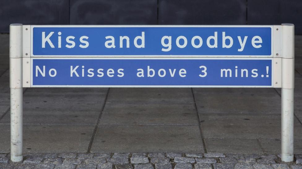 A blue sign with white writing at the drop off area at Aalborg airport in Denmark. It says kiss and goodbye! No kisses above 3 mins!.