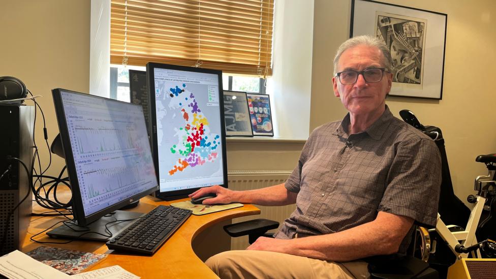 A man in his sixties wearing reading glasses, a brown short-sleeved shirt and lighter brown trousers sits behind his desk with several computer screens in front of him. One on the right displays a map of England and Wales with coloured dots on it. The screen of the left computer is just visible and shows a couple of graphs. In the background is a window, radiator, framed awards and a bike machine. 