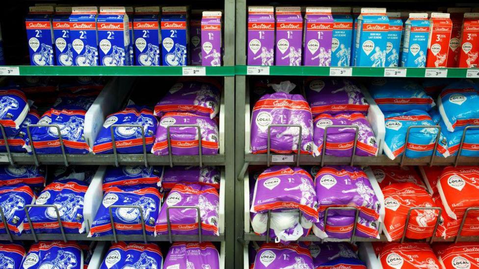 Shelves of milk, with milk cartons on a top shelf and bags of milk on lower shelves, with different colour packaging - blue, purple or red - representing the fat percentage. 