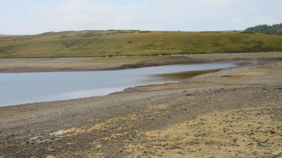 Llyn Aled Isaf in Conwy. The banks of the lake are dried up, the nearest bank is sandy with pebbles on, the bank in the distance is green, with a hill rising up behind it.