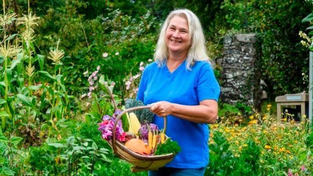 Stephanie Hafferty standing in her garden holding a wicker basket of homegrown vegetables. She is wearing a blue top and jeans, and has long white hair.