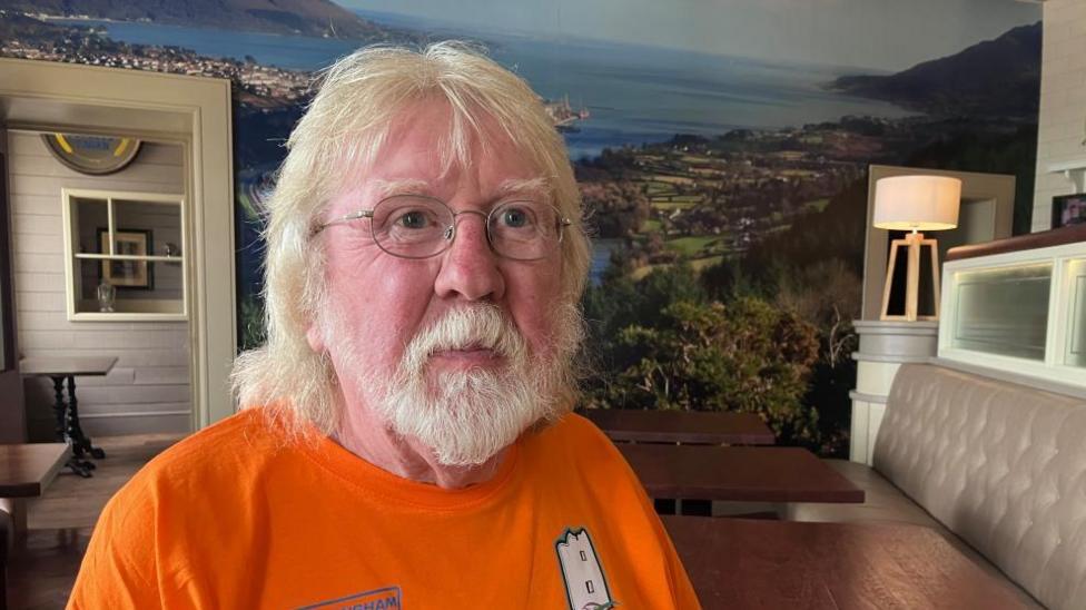 Quinn is wearing a bright orange t-shirt and has long white hair and a white beard.