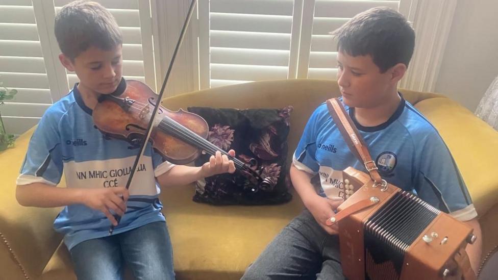 Two young boys are sitting on a yellow sofa. They are wearing light blue and white jersey-style tops. On the left the boy is playing a fiddle and the boy on the right is playing an accordion. 
