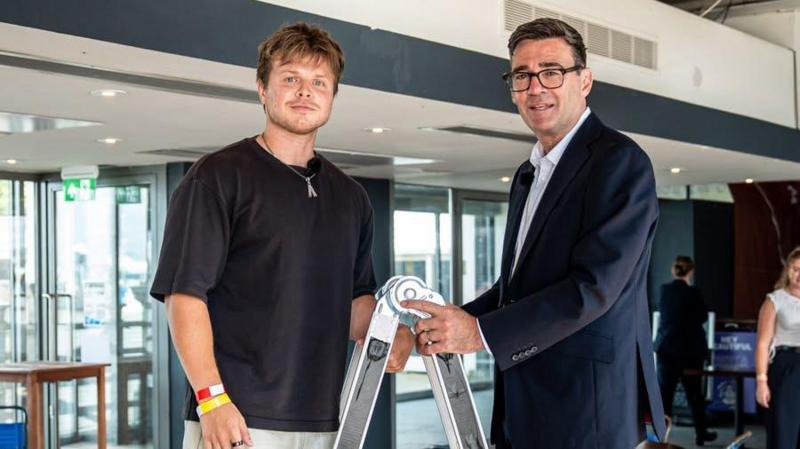 Andy Burnham, in a suit jacket and open necked shirt, stands at the top of a step ladder in an open plan office, opposite Max Klymenko, a young man with a light beard, wearing a black T-shirt