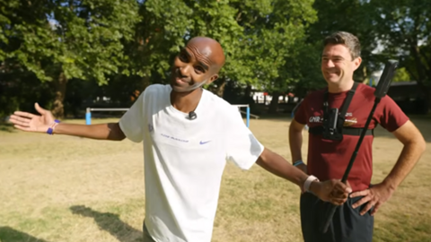 Sir Mo Farah smiles and spreads his arms wide, as he holds a selfie stick in a park with dry, parched grass and trees. He is wearing a white Nike T-shirt. Andy Burnham is standing behind him with his hands on his hips. He is wearing a red T-shirt and jogging bottoms.