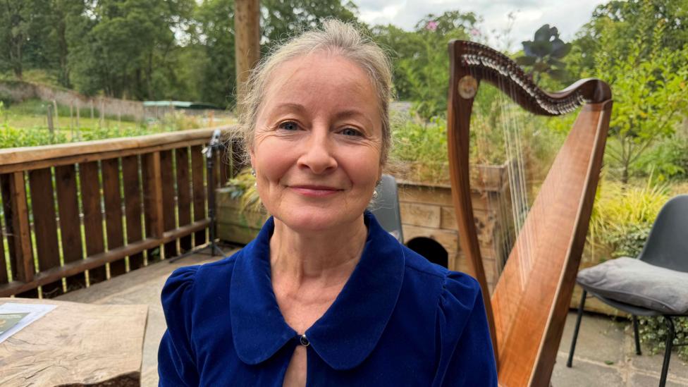 A woman wearing a dark-blue blazer smiles at the camera. She has grey hair and is standing in front of a harp. Behind her are trees.