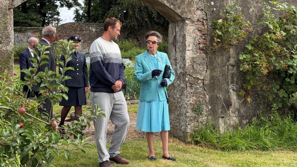 Princess Anne wearing a teal outfit. She is standing next to a man with short brown hair wearing a grey jumper. Princess Anne is wearing sunglasses and gloves. Behind them are three people. Princess Anne and the man next to her are standing on a patch of grass in front of a stone archway. There are ushes around them.