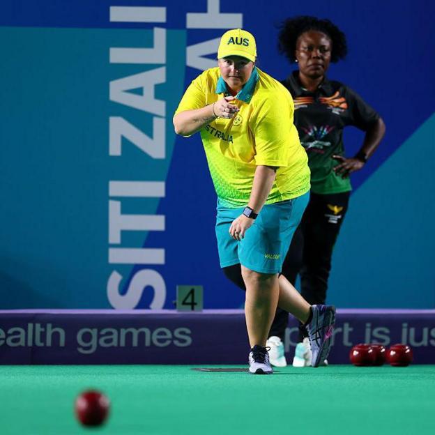 Dawn Hayman, a bowler, looking at her shot while standing on an indoor rink. She is wearing a yellow shirt and green shorts. 