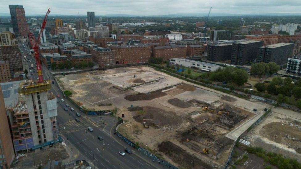 An aerial photograph taken with a drone shows construction on a large, brownfield site in Manchester. Tower blocks can be seen in the distance, smaller office blocks are nearby and there is a multi-lane road running along the left hand side. A red crane stands on the left hand side of the picture.