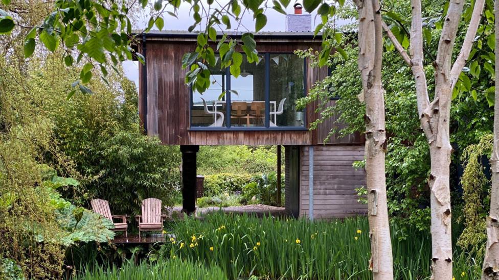 A timber house on stills above a lake. There's greenery all around and the house has huge windows to overlook the views. Inside, a table and chairs can be seen by the window.