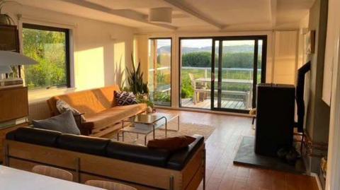 A large living room with 60s style sofas in an L shape, facing a wood burner. The sun is beaming in through the open French doors overlooking the estuary.
