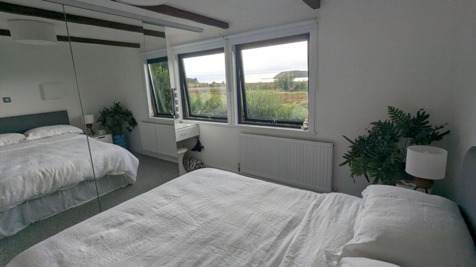 A clean and crisp bedroom with a double bed with white sheets on it. There's a green plant in the corner and large windows with views of the countryside.