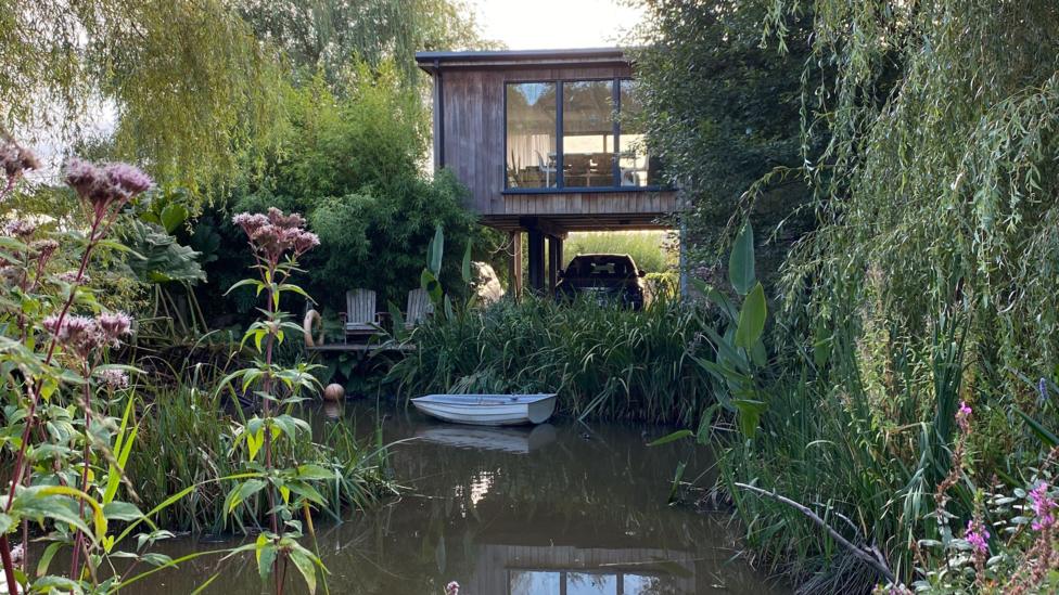 A timber house on stills above a lake. There's greenery all around and the house has huge windows to overlook the views. There's a little boat on the water.