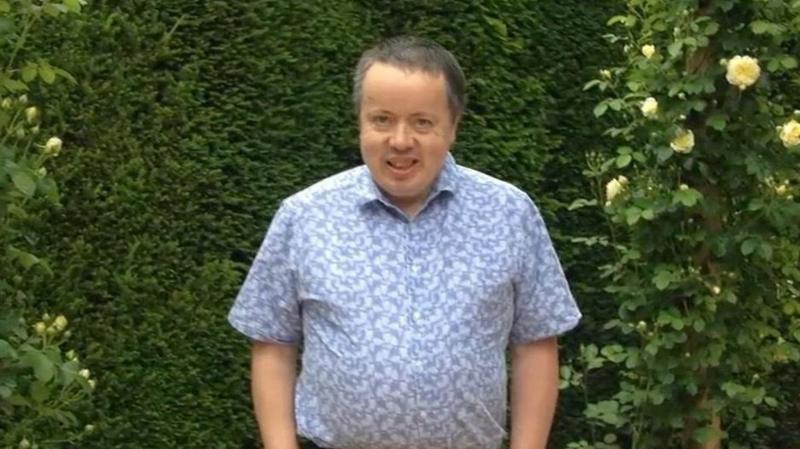 A man with grey hair smiling at the camera in front of green bushes. He is wearing a short sleeved blue patterned shirt.