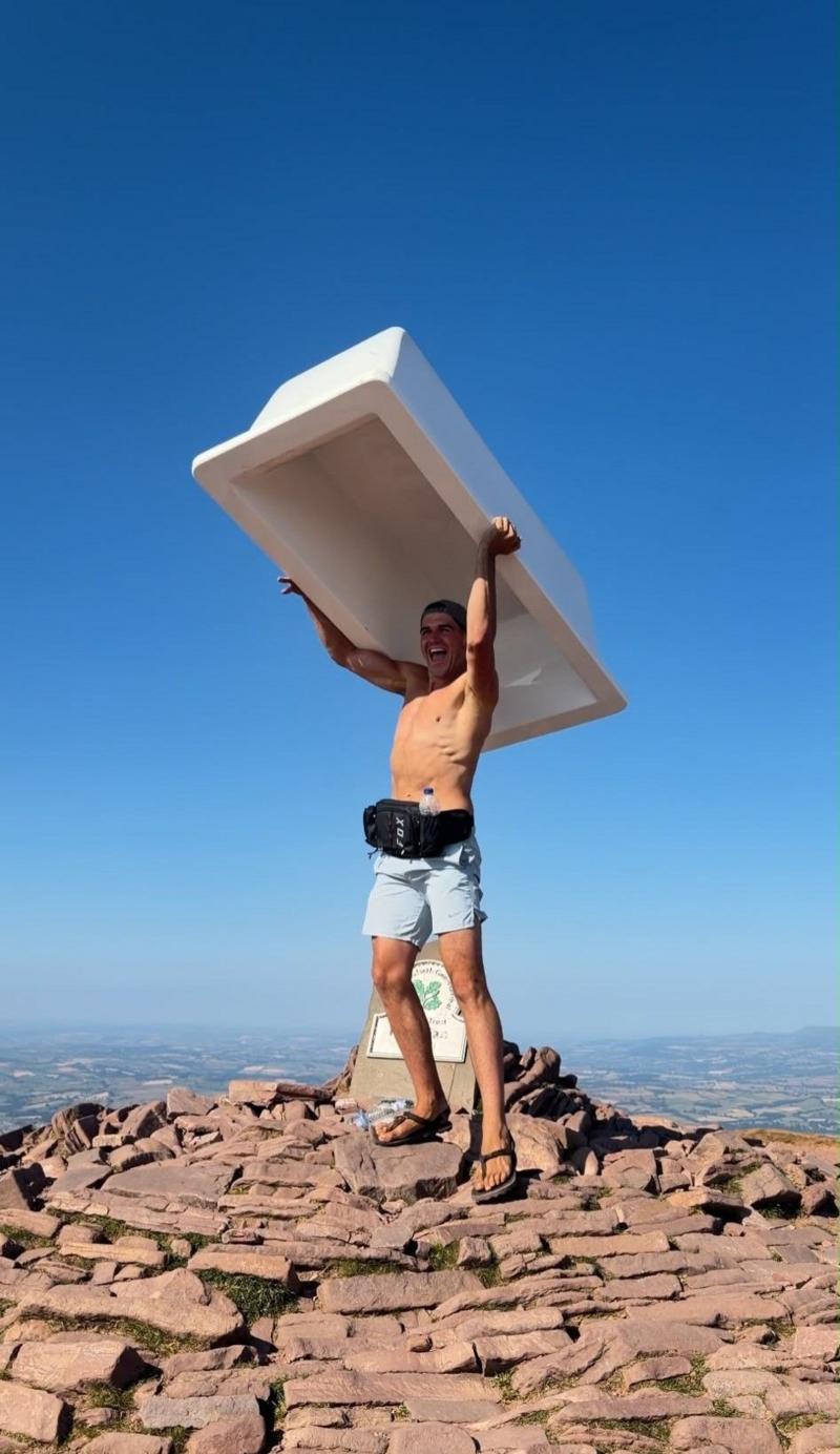 A shirtless man is lifting a bath tub above his head with his arms while standing on stones and rocks on a mountain summit