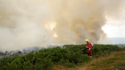 Firefighter pictured on the mountain wearing a yellow helmet and red suit, spraying water onto bracken as smoke billows behind him.