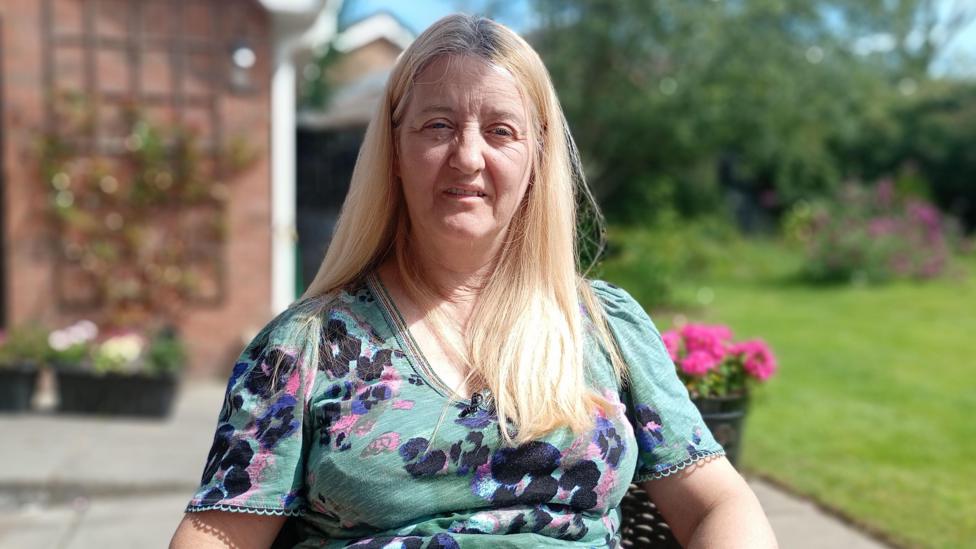 Sharon has blond hair and is sitting in a garden. She's wearing a floral top. Flowers are in the background as well as a paved patio, a grassy lawn and some foliage.