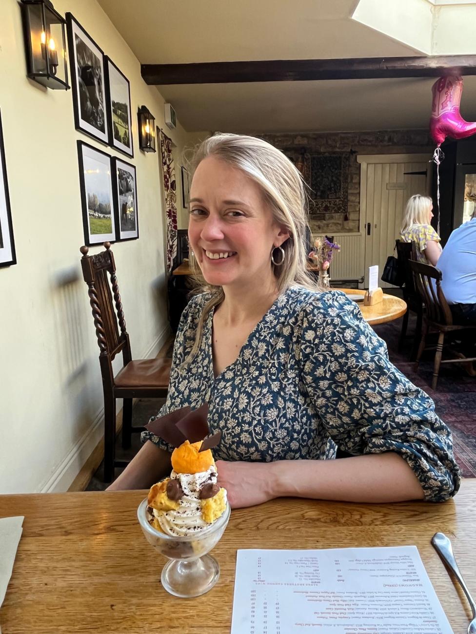 Photo of young woman sitting at a table in a restaurant. She has blonde hair tied back and is wearing a floral blouse. In front of her sits a sundae ice cream dessert.