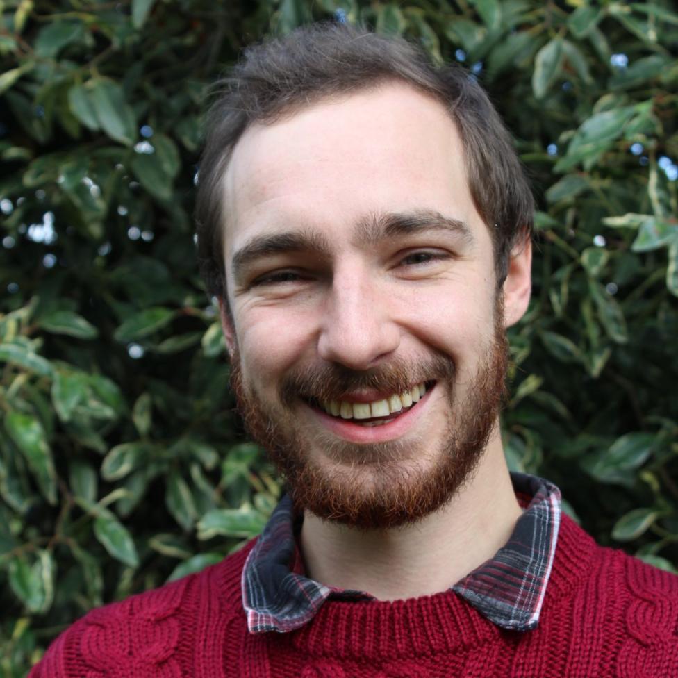 Headshot of young man with short brown hair and beard. He wears a checked shirt under a red jumper and is grinning at the camera.