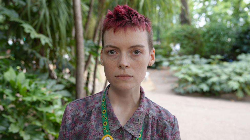 A person with short spiky hair looking at the camera. They are wearing a floral shirt and a green lanyard with sunflowers on it. Behind them is a path and greenery.
