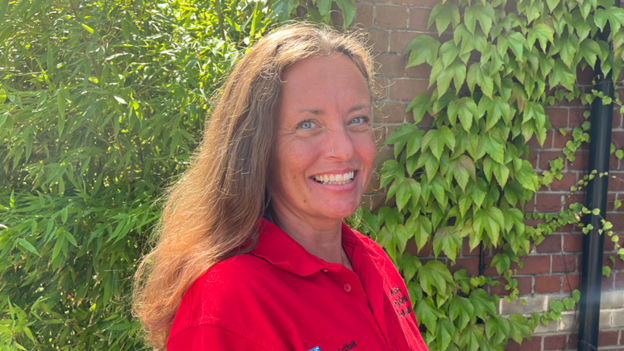 Vicki Bailey is standing in front of a wall covered in green leaves. She has shoulder-length brown hair and is wearing a red polo shirt.