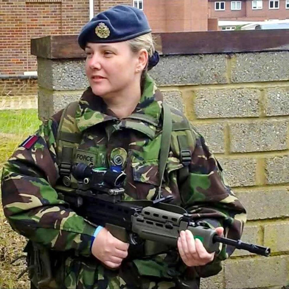 A woman is wearing army fatigues and is holding a very-realistic looking training gun. She is wearing a blue beret and has her blonde hair tied back.