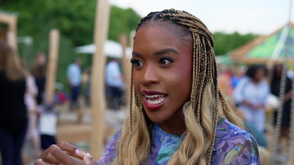 Amber Ogunsanya-Williams has dark and blonde braided hair. Behind her are kids playing in the chalk stream area.