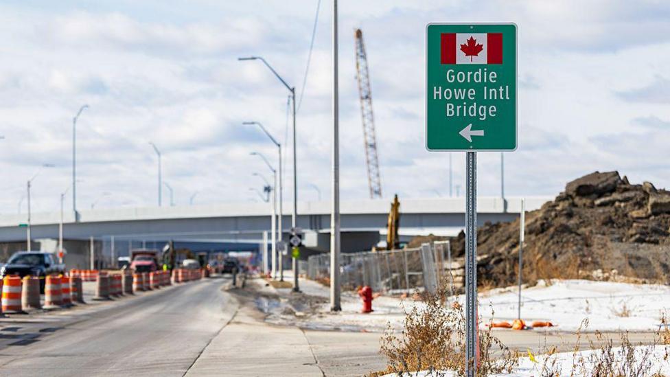 A sign for the Gordie Howe International Bridge, with a Canadian flag and an arrow pointing left, sits on the side of a highway. 