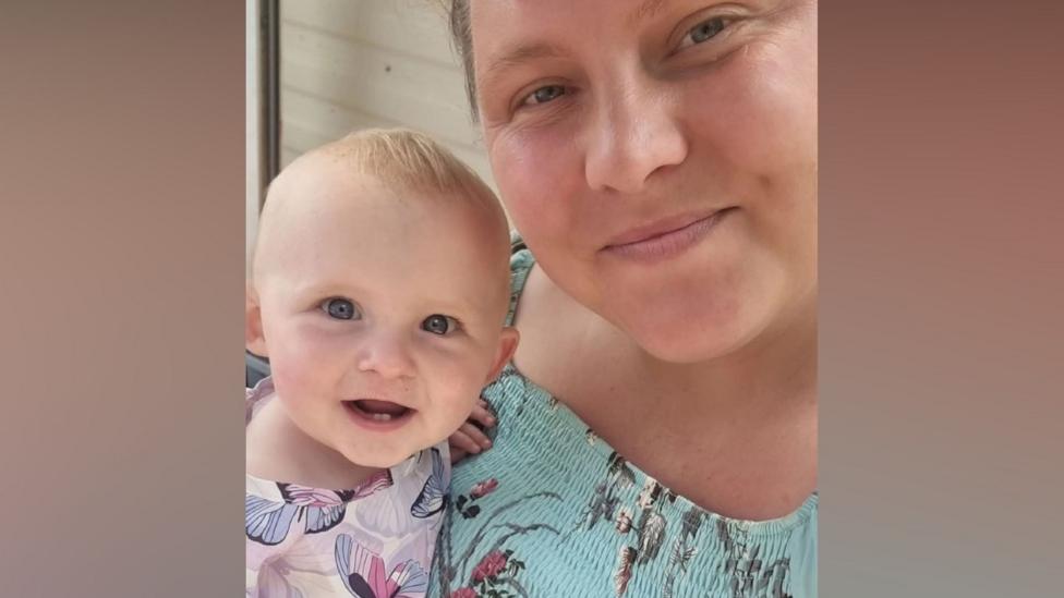 A mum poses for a selfie with her baby daughter. The mum has brown hair which is tied back and is wearing a light blue top with a floral pattern on. Her baby is smiling, has very short blonde hair and is wearing a pink, purple and white dress with butterflies.