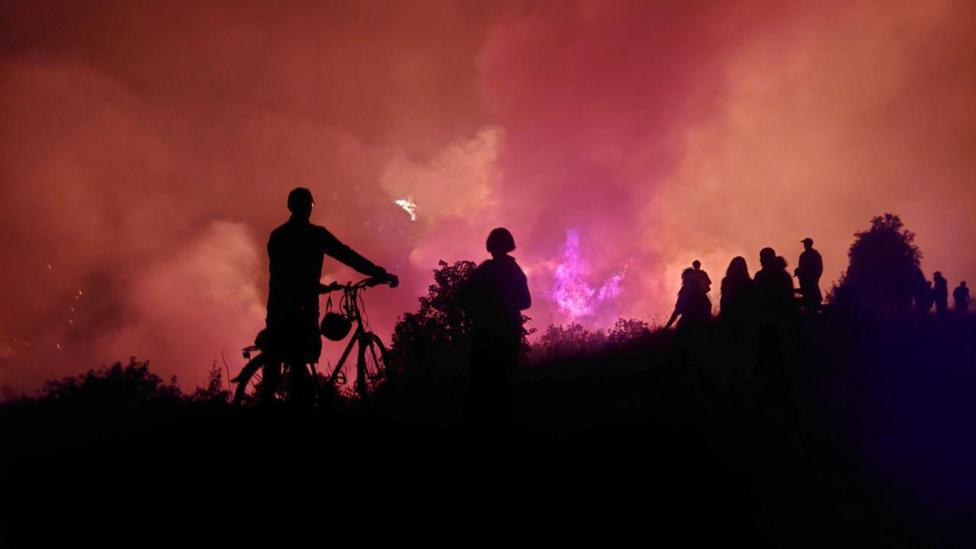 Onlookers in silhouette look on as a fire rage in the background on a hill
