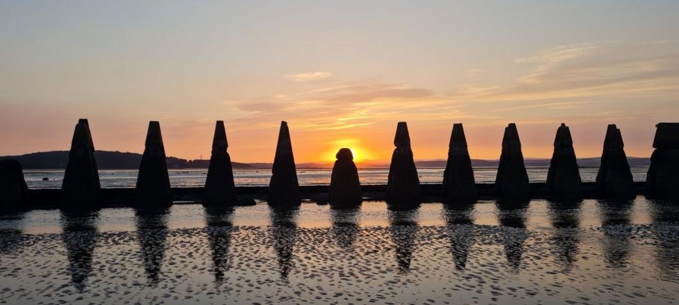 A row of concrete pylons on a beach, with the sun setting behind the smaller middle pylon