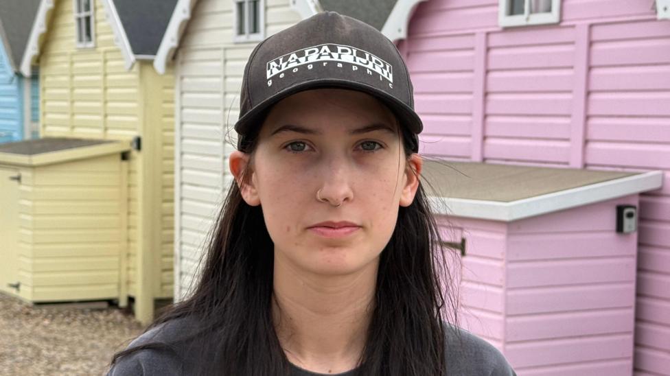 Leonie Powling stands on a stony beach, in front of multicoloured beach huts. She wears a dark-coloured jumper and black baseball cap.