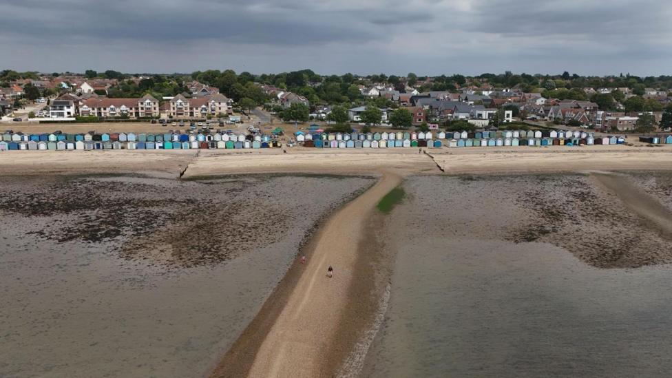 A drone image showing a long row of beach huts and the sandy beach in front of them. The water has been pictured at low tide. There is a long sandbank that splits the middle of the water.