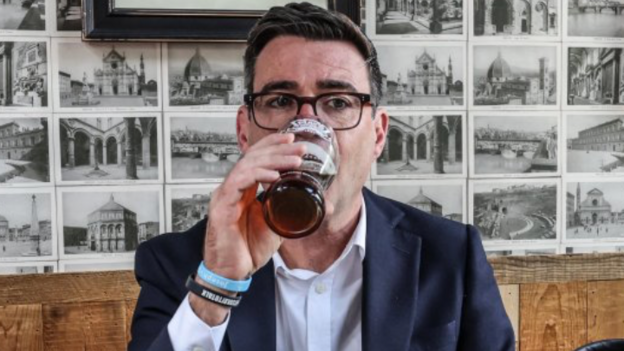 The new prime minister wears a suit in a pub and drinks a beer from a pint glass.