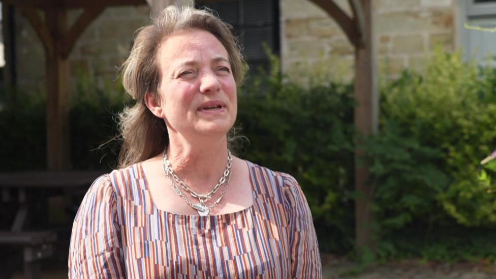 A woman wearing a colorful stripy blouse and silver necklaces speaks to a journalist while being filmed on camera in the sunshine.