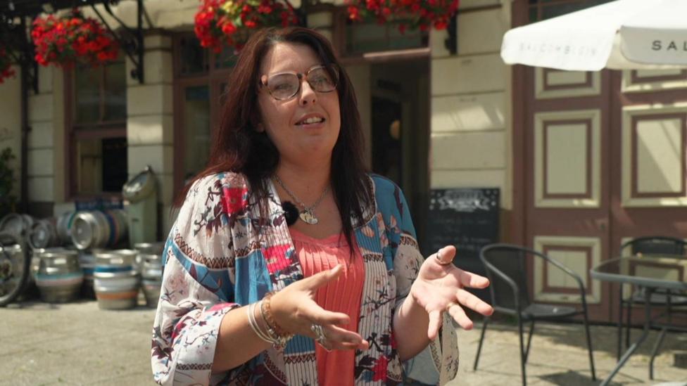 A woman wearing an orange top under a colorful jacket and glasses stands outside a pub while speaking to the camera.