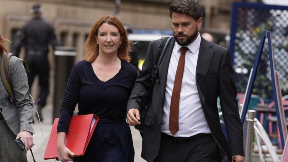 Louise Haigh is walking down the street next to an aide. She is carrying a large, scarlet government folder and wears a smart black top and black trousers. Her hair is ginger and shoulder length, and she wears it loose, along with a pair of small earrings.