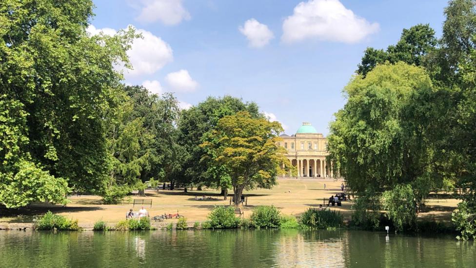 Pitville Park in Cheltenham, Gloucestershire. There is a large beige building with a green dome roof in the distance. There is a body of water and trees dotted around the park. People can be seen sat on benches in the sun and shade.