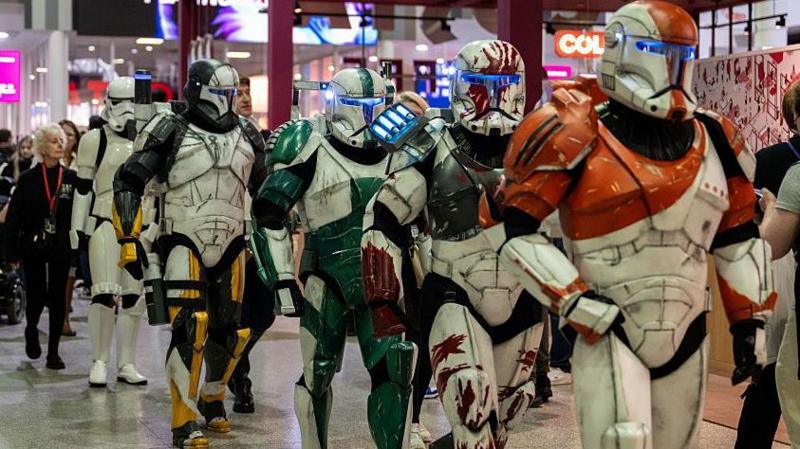 Cosplayers dressed as Clonetroopers and Stormtroopers from the Star Wars films walking across the floor of a convention. They are wearing white armour, mixed with other colours, such as orange or green.