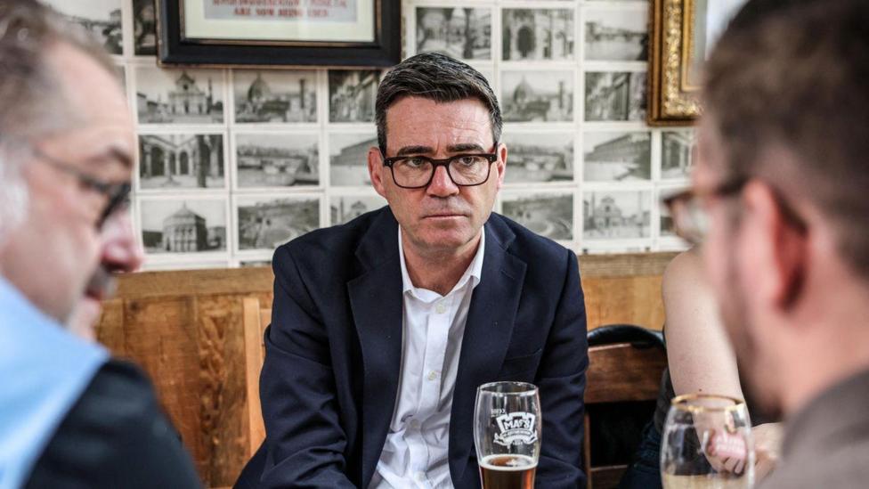 Andy Burnham has dark hair and is wearing dark glasses. He is wearing a white shirt with a dark suit jacket and is sitting at a table with a pint in front of him. He is facing two men who are talking