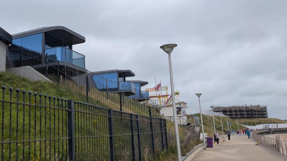 Three cabins are visible on a grassy bank overlooking a beach walkway. 