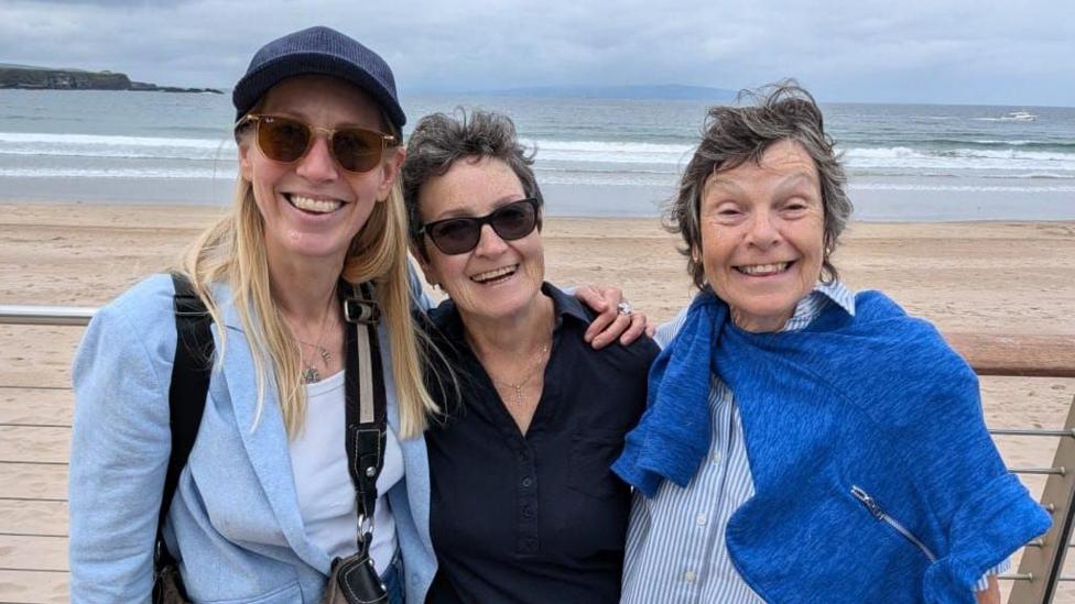 Helen Quinn is wearing a blue striped shirt and navy blue shawl. She has short brown hair and is standing near her two friends who are both wearing sunglasses.