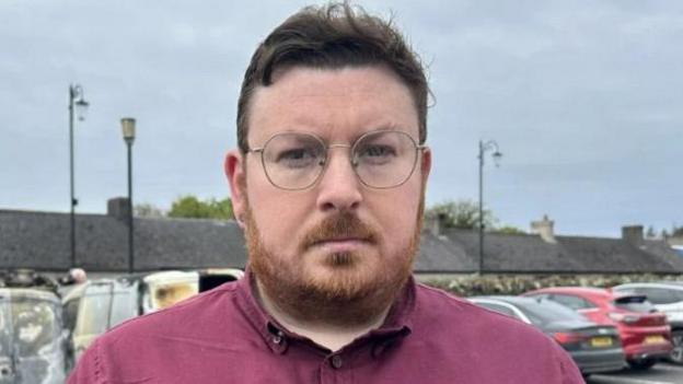  Richard Brown Stewart has short brown hair and a beard. He is wearing a purple shirt. He has circular glasses and is standing in a car park. 