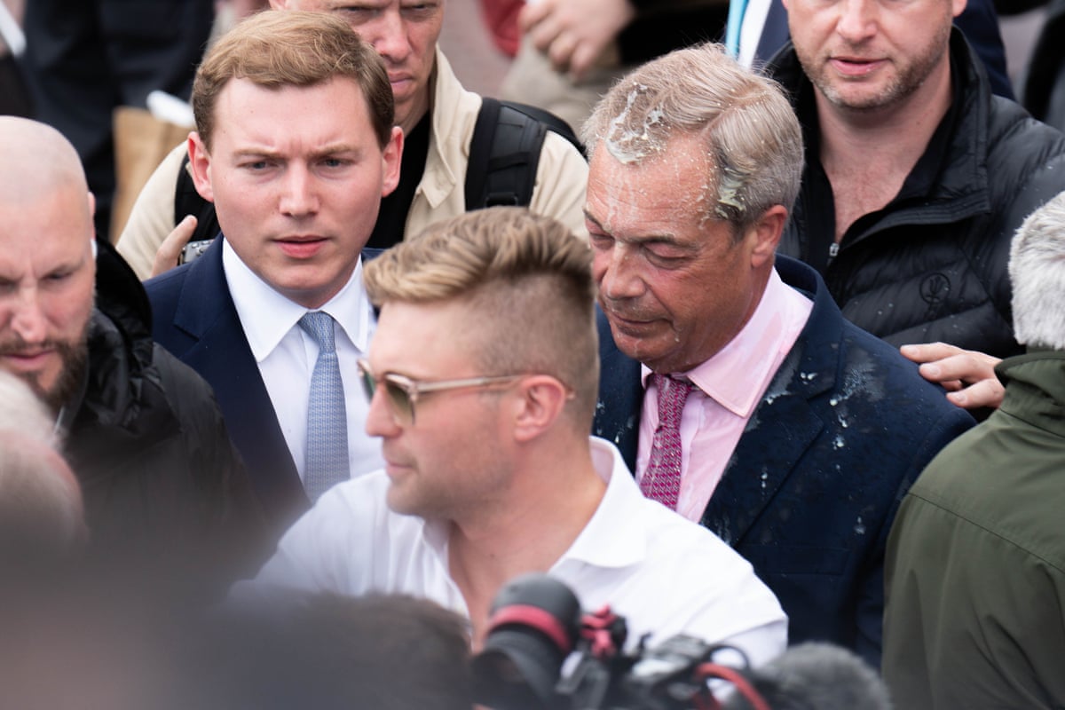 Nigel Farage, with splatters of milkshake on his face and head, in a crowd next to George Cottrell.