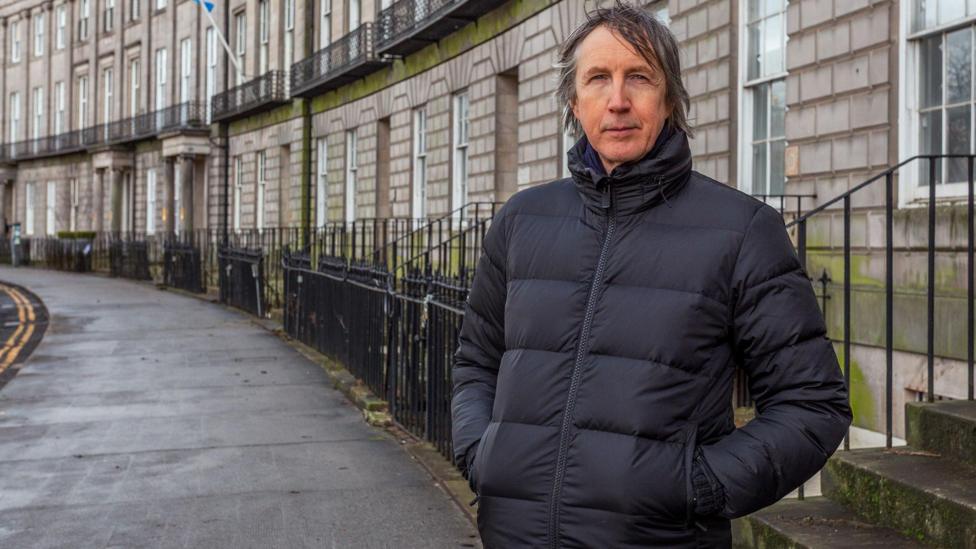 Philip Flockhart has long hair and is wearing a blue puffer jacket. He is standing on front of the crescent of Georgian houses.