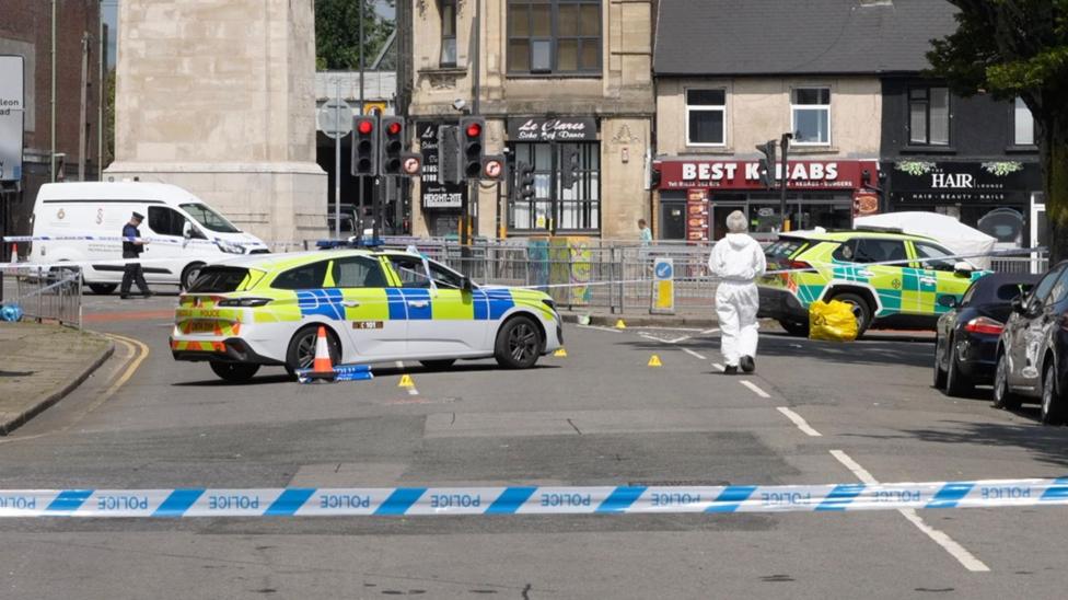 A police cordon can be seen in the forefront. Behind it is a police car parked on a stretch of road, with yellow evidence cones on the ground. A forensic officer wearing a white overall can be seen walking away from the camera.
