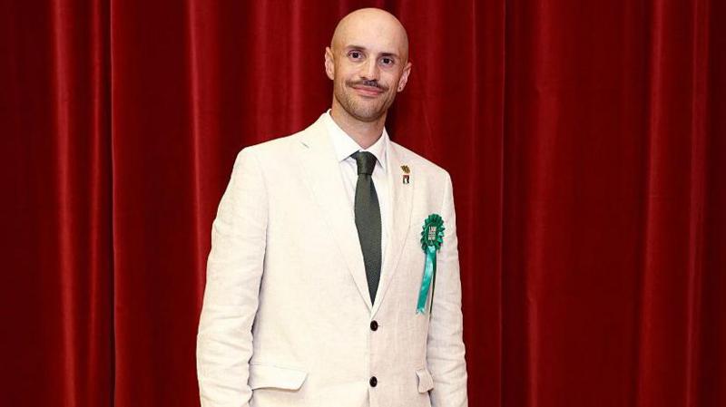 A man in a cream linen suit, wearing a green rosette and green tie, stands in front of a red theatre curtain.