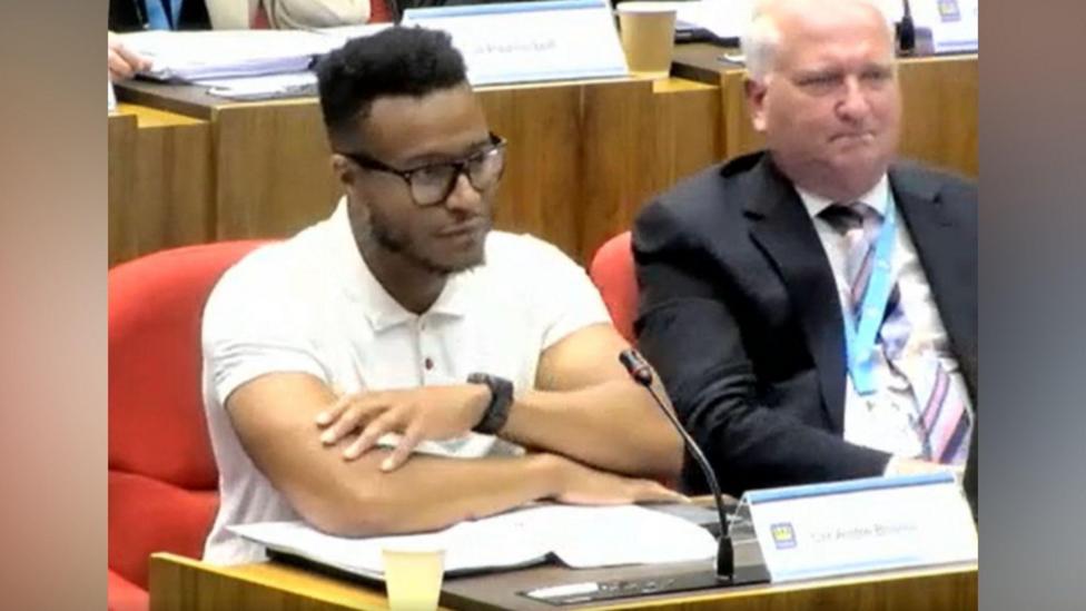 A screen shot of a council meeting - a councillor sits at a desk and is wearing a white T-shirt and glasses sits with his arms folded over some paperwork. There is a sign in front of him that reads Cllr Andre Bourne. Beside sits another man in a dark suit, white shirts and striped tie. There are other people sat at similar desks in the background.